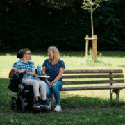 Auf einer Parkbank sitzt eine junge Frau und unterhält sich fröhlich mit einem jungen Mann, der im Rollstuhl direkt neben der Bank sitzt.