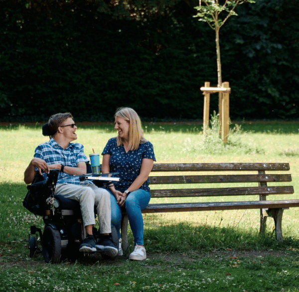 Auf einer Parkbank sitzt eine junge Frau und unterhält sich fröhlich mit einem jungen Mann, der im Rollstuhl direkt neben der Bank sitzt.