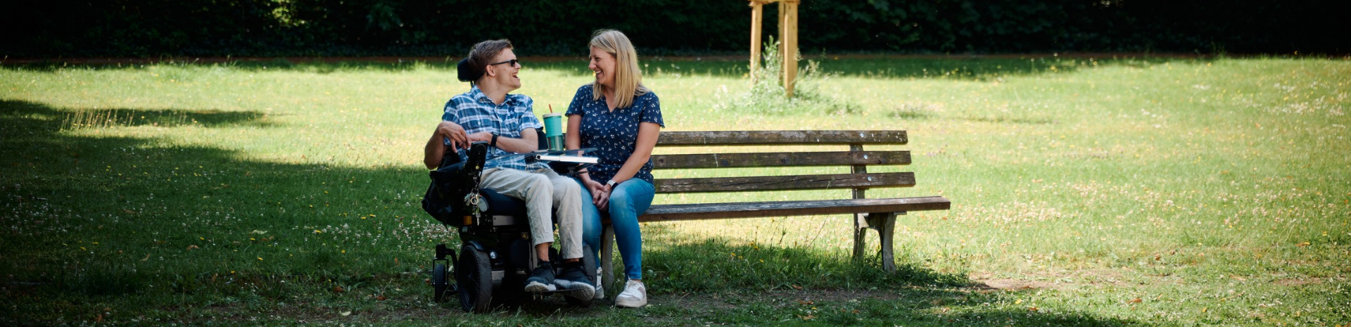 Auf einer Parkbank sitzt eine junge Frau und unterhält sich fröhlich mit einem jungen Mann, der im Rollstuhl direkt neben der Bank sitzt.